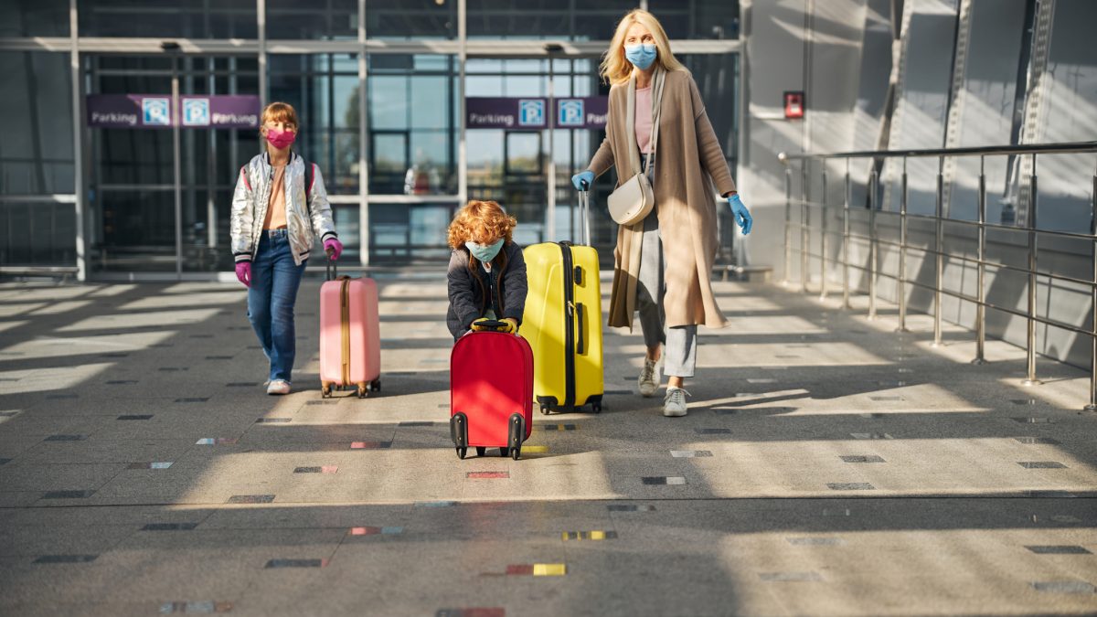 Eine Frau läuft mit zwei Kindern an einem Flughafengate entlang. Sie haben Koffer bei sich und tragen Mundnasenschutz.