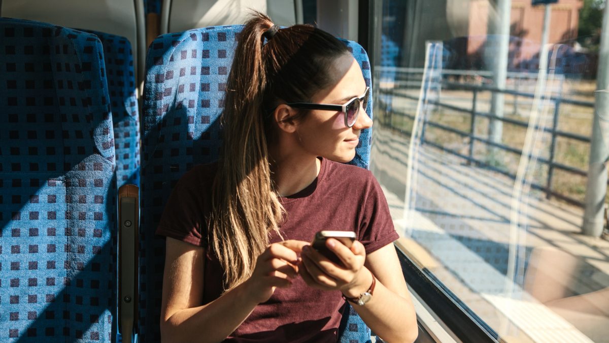 Eine junge Frau sitzt in einem Zug und sieht aus dem Fenster. In der Hand hält sie ein Mobilfunkgerät.