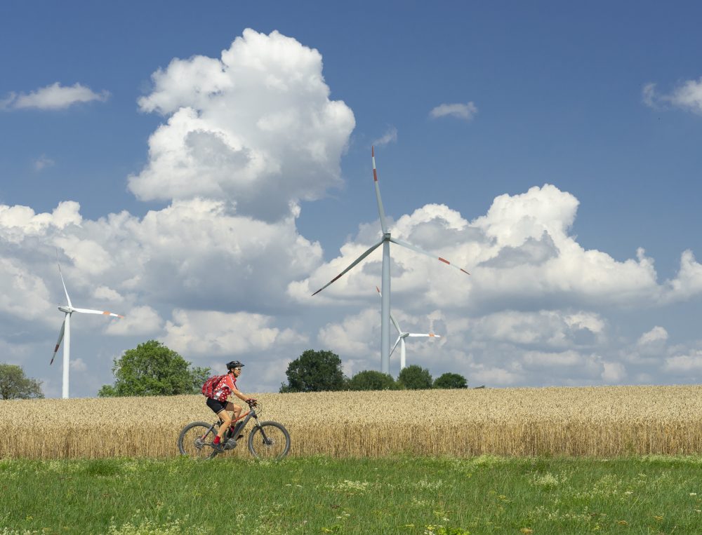 Eine Person fährt auf einem Fahrrad durch die Natur. Im Hintergrund sind ein Kornfeld und Windräder zu sehen.