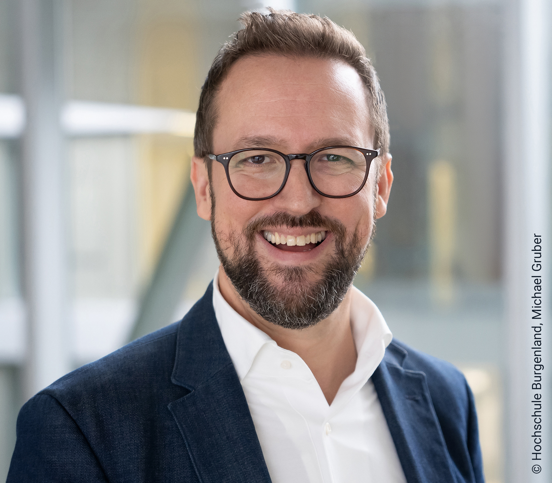Ein Mann mit Bart und Brille, der ein weißes Hemd und einen blauen Blazer trägt.