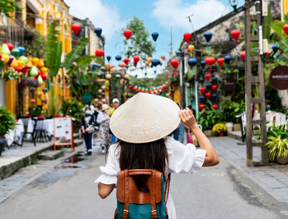 Eine Person mit Rucksack und kegelf&ouml;rmigem Hut geht eine farbenfrohe Stra&szlig;e entlang, die mit h&auml;ngenden Laternen geschm&uuml;ckt ist, in einer asiatischen Stadt mit gelben Geb&auml;uden und viel Gr&uuml;n auf beiden Seiten.