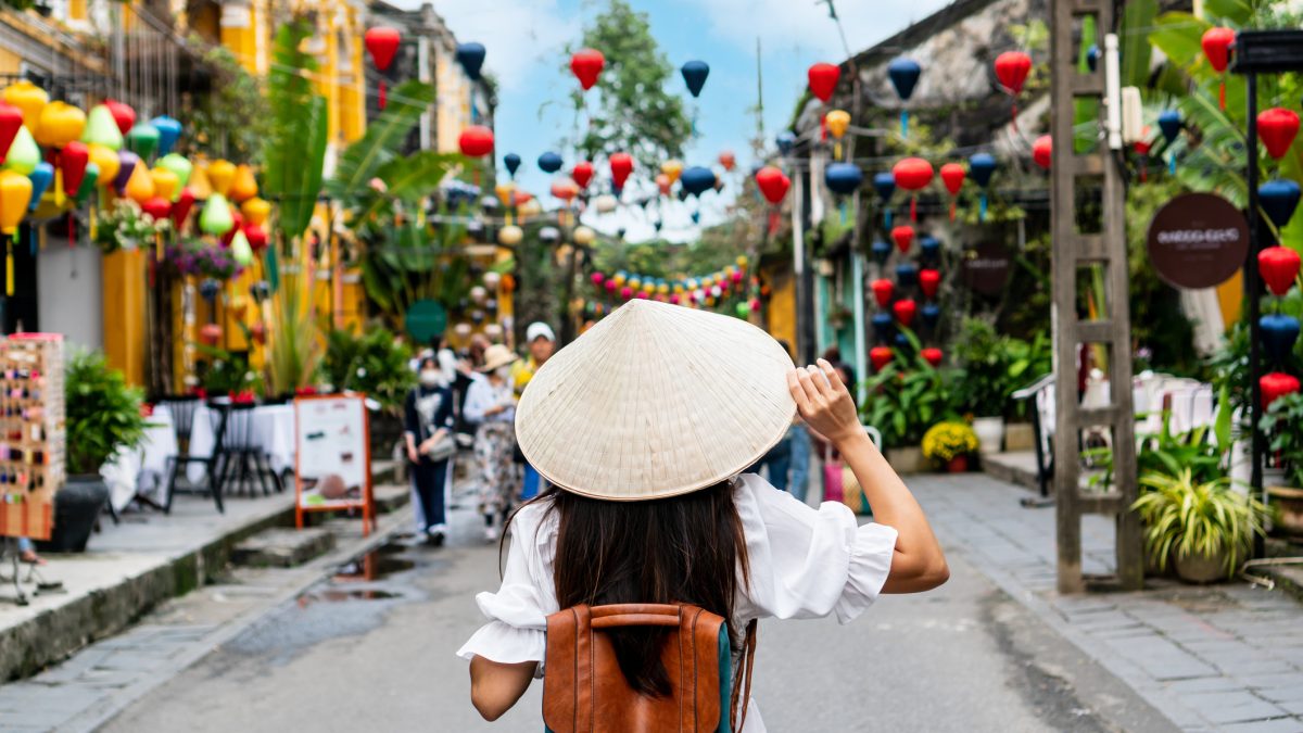 Eine Person mit Rucksack und kegelförmigem Hut geht eine farbenfrohe Straße entlang, die mit hängenden Laternen geschmückt ist, in einer asiatischen Stadt mit gelben Gebäuden und viel Grün auf beiden Seiten.